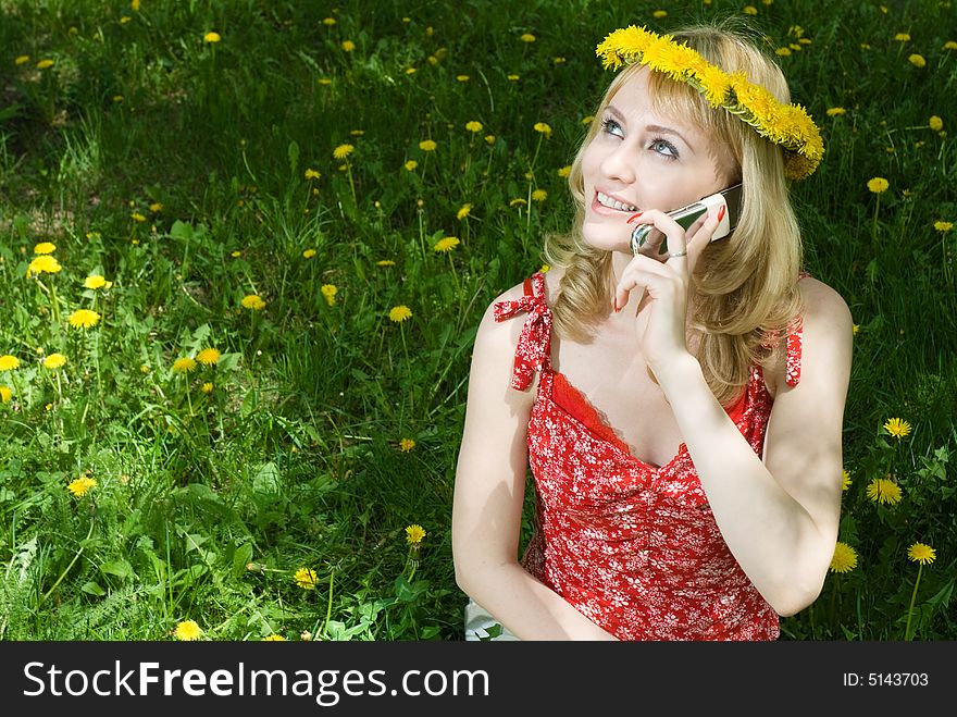 Young beautiful blond hair woman in garland talking on the mobile phone, sitting on the grass among dandelions. Young beautiful blond hair woman in garland talking on the mobile phone, sitting on the grass among dandelions