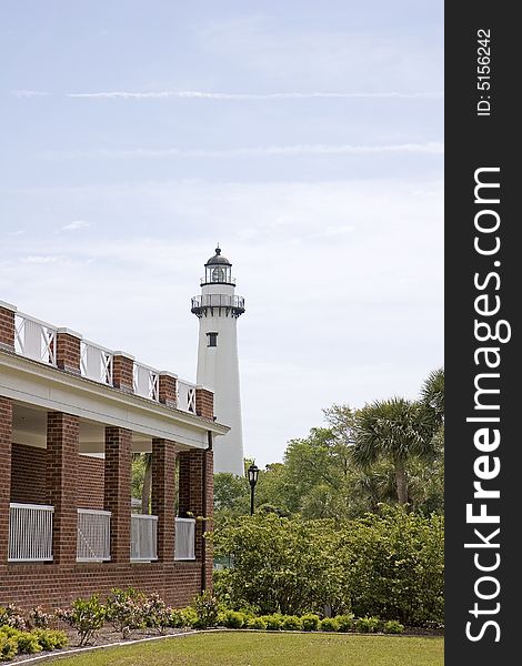 A lighthouse on the horizon past a brick building and trees. A lighthouse on the horizon past a brick building and trees