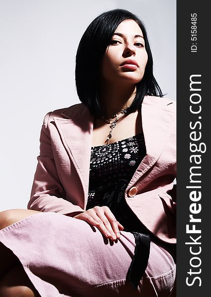 A portrait about an attractive trendy lady with black hair who is sitting on a chair, she is looking ahead and she has a glamorous look. She is wearing a nice dress, a pink skirt, a necklace and a pink stylish coat. A portrait about an attractive trendy lady with black hair who is sitting on a chair, she is looking ahead and she has a glamorous look. She is wearing a nice dress, a pink skirt, a necklace and a pink stylish coat.