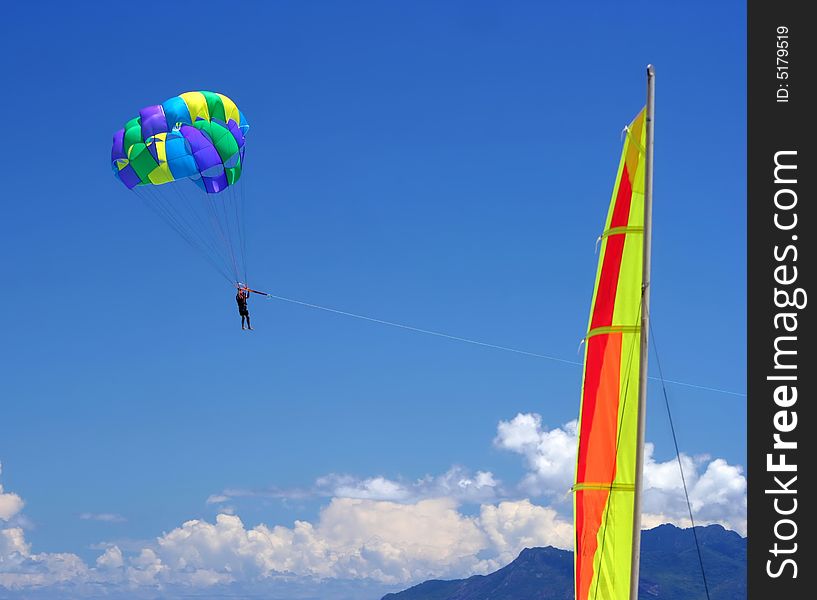 Para-sailing over sea. Colorful parachute with parachuter is on the blue sky, above the white clouds & mountain. The colorful sail is in front of it . Para-sailing over sea. Colorful parachute with parachuter is on the blue sky, above the white clouds & mountain. The colorful sail is in front of it .