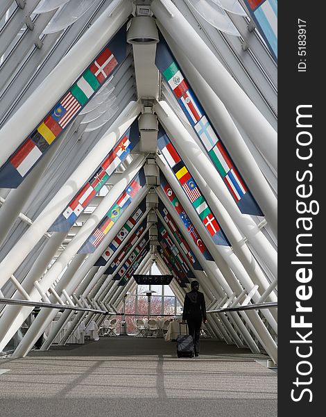 Businesswoman walking through a walkway / skybridge pulling her luggage behind her. The walkway is lined with flags. Vertically framed shot with the woman walking away from the camera. Businesswoman walking through a walkway / skybridge pulling her luggage behind her. The walkway is lined with flags. Vertically framed shot with the woman walking away from the camera.