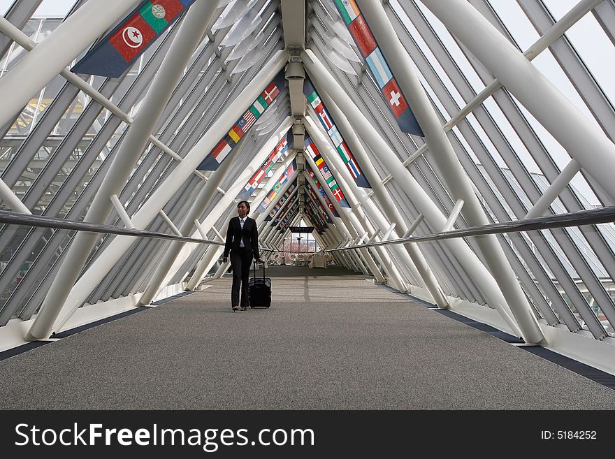 Businesswoman walking through a walkway / skybridge pulling her luggage behind her. The walkway is lined with flags. Horizontally framed shot with the woman walking towards the camera. Businesswoman walking through a walkway / skybridge pulling her luggage behind her. The walkway is lined with flags. Horizontally framed shot with the woman walking towards the camera.