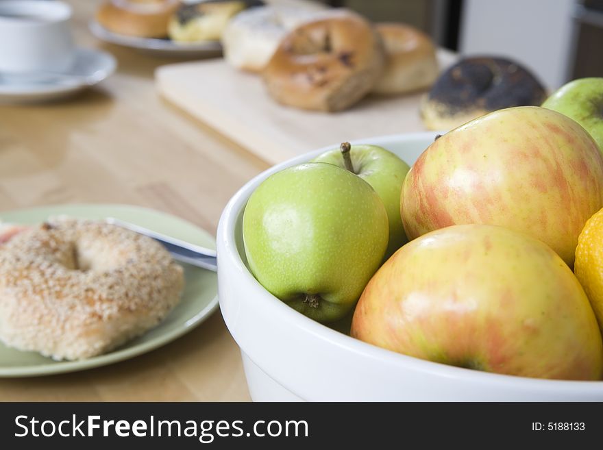 Breakfast foods on top a kitchen counter.  A bowl of apple, bagels on a plate and coffee cup in the background. Breakfast foods on top a kitchen counter.  A bowl of apple, bagels on a plate and coffee cup in the background.