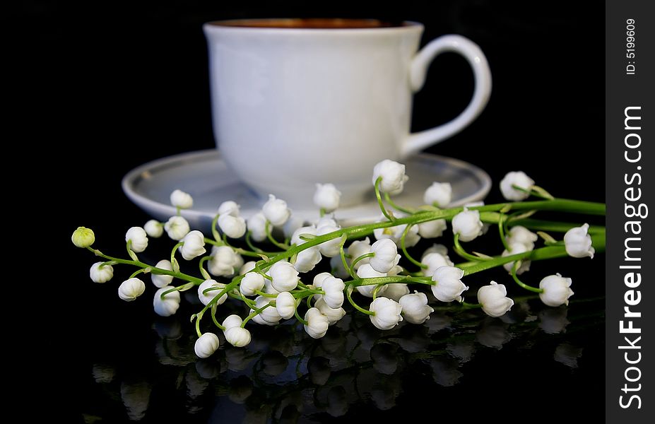 Flowers of a llies of the valley and white cup of black coffee on a black background. Flowers of a llies of the valley and white cup of black coffee on a black background