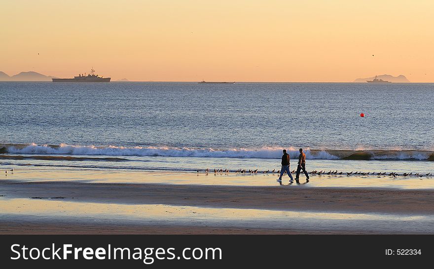 Two friends take a stroll on Southern California beach at sunset. Navy ships and Coronado Islands (Mexico) in the background. (Coronado, CA). Two friends take a stroll on Southern California beach at sunset. Navy ships and Coronado Islands (Mexico) in the background. (Coronado, CA)