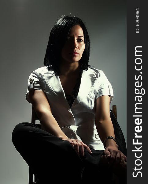 A low-key portrait about an attractive trendy girl with black hair who is sitting on a chair, she is looking ahead and she has a charming look. She is wearing black pants and a white stylish coat. A low-key portrait about an attractive trendy girl with black hair who is sitting on a chair, she is looking ahead and she has a charming look. She is wearing black pants and a white stylish coat.