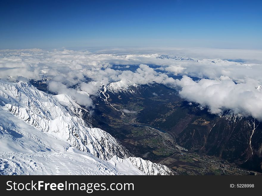 Spring valley Chamonix between high snow mountains occupied by light layer of white clouds and blue sky, view from above. Spring valley Chamonix between high snow mountains occupied by light layer of white clouds and blue sky, view from above