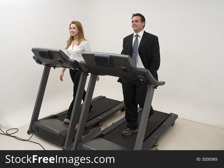 Two businesspeople (male and female) running on a treadmill and looking at each other facing forwards. Two businesspeople (male and female) running on a treadmill and looking at each other facing forwards