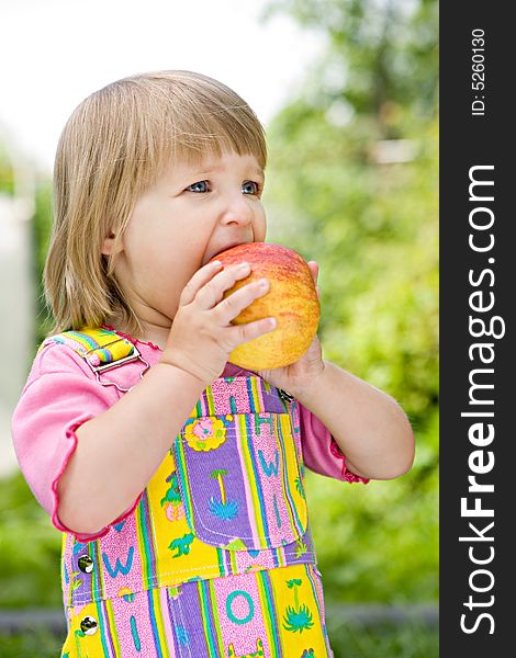 Little girl in a multi-coloured dress bites off an apple in the solar afternoon. Little girl in a multi-coloured dress bites off an apple in the solar afternoon