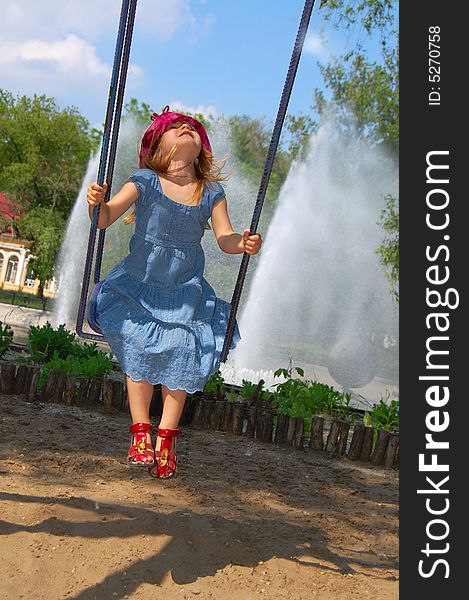 Little pretty swinging girl on the background of blue sky, trees and fountain. Little pretty swinging girl on the background of blue sky, trees and fountain