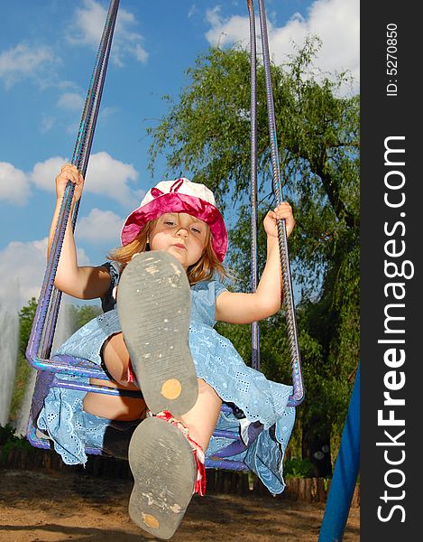Little pretty swinging girl on the background of blue sky, trees and fountain