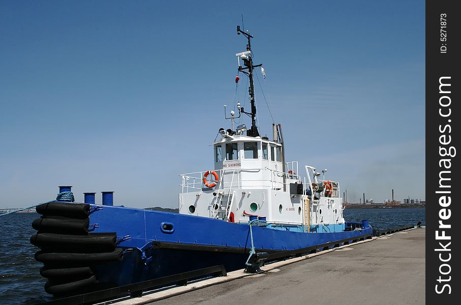 A white and blue tug boat is docked at the pier with industry in the background. A white and blue tug boat is docked at the pier with industry in the background.