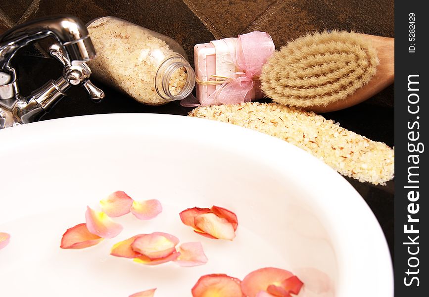 Bath salt, soap and a wooden brush laying around a white ceramic sink with rose petals floating in the water. Bath salt, soap and a wooden brush laying around a white ceramic sink with rose petals floating in the water