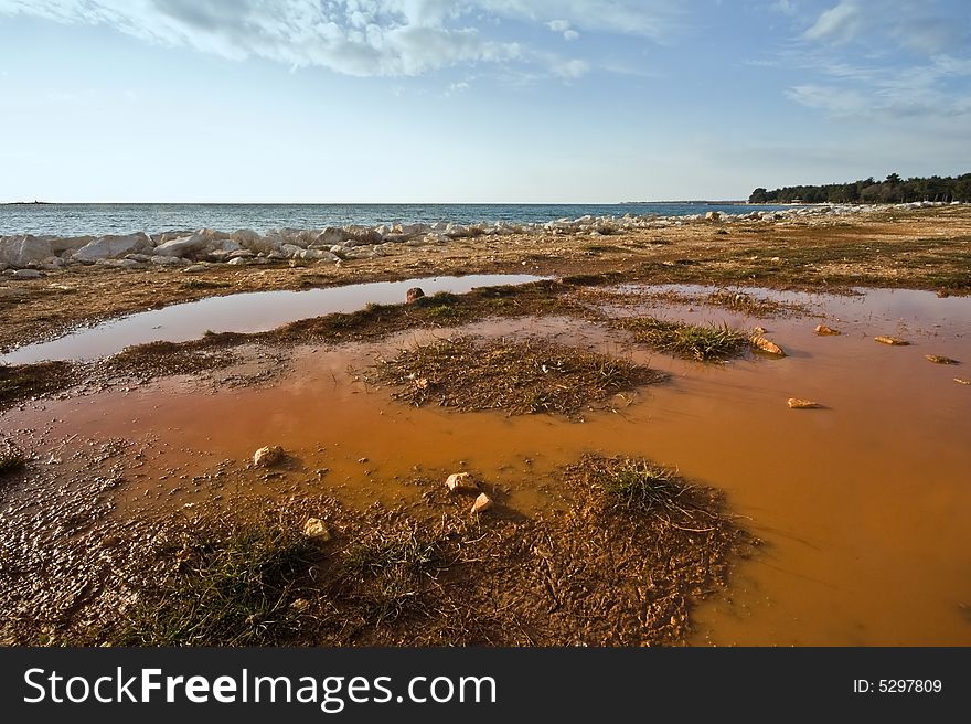 Pond on red dirt near sea with cloudy sky