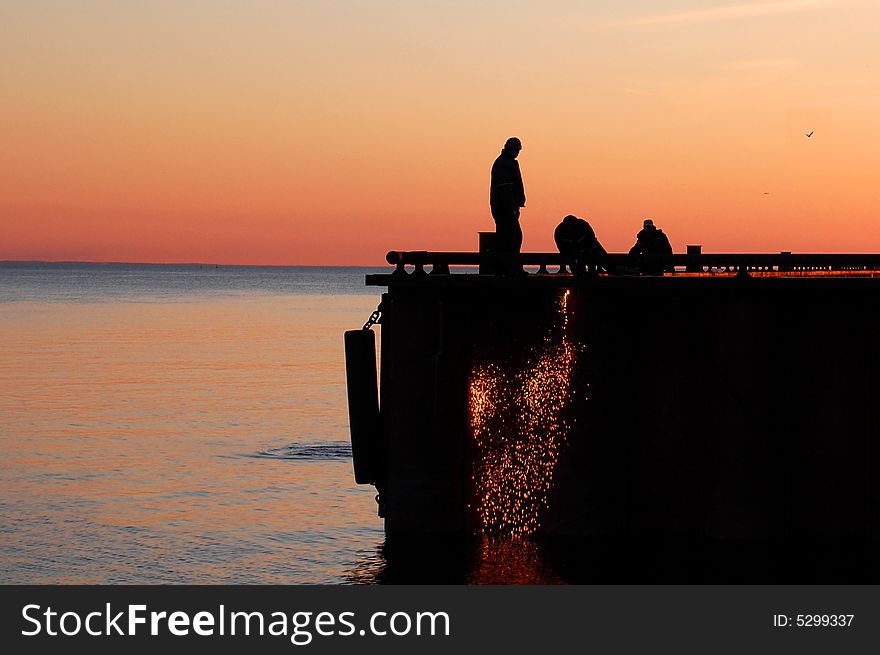 Silhouette of three workers in a port fixing a moorage, at red light. A flow of sparks is falling to the water. Silhouette of three workers in a port fixing a moorage, at red light. A flow of sparks is falling to the water.