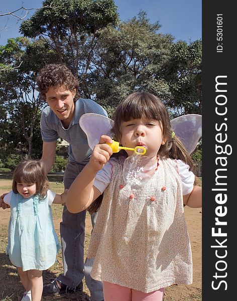 Cute young girl blowing bubbles while her younger sister and father look on. Vertically framed shot.