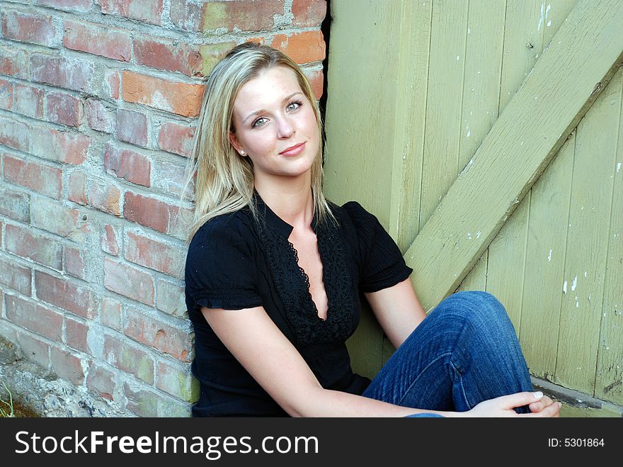 Attractive smiling woman sitting, with one knee pulled up, sitting next to a brick building and green colored wood carraige door. Attractive smiling woman sitting, with one knee pulled up, sitting next to a brick building and green colored wood carraige door.