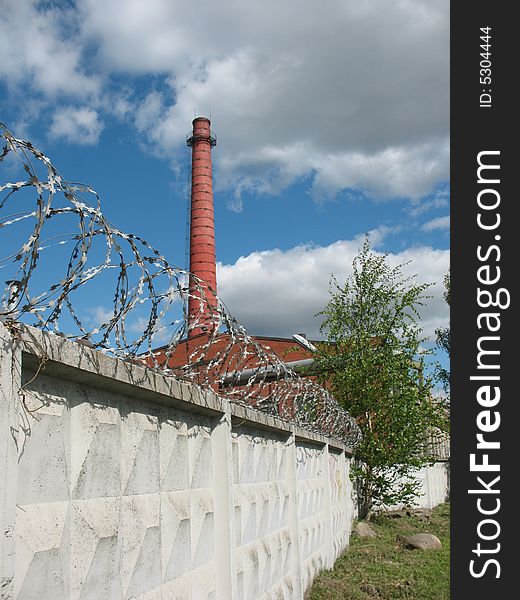 A secured industrial zone with concrete fence, barbed wire and brick chimney-stalk on the blue sky background.