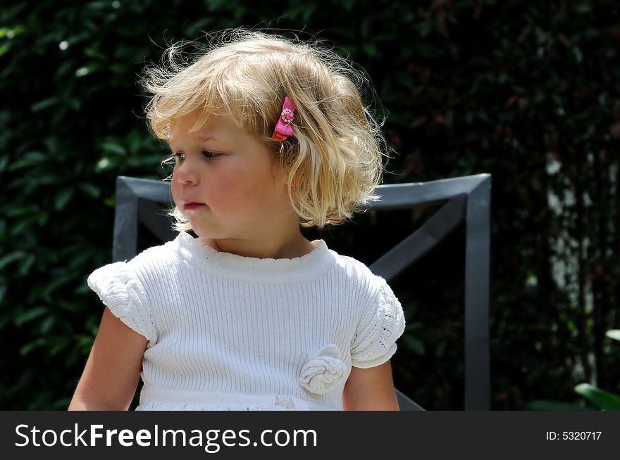 Little girl sitting outside in a garden on a chair, gazing pensively off to the side. Little girl sitting outside in a garden on a chair, gazing pensively off to the side.