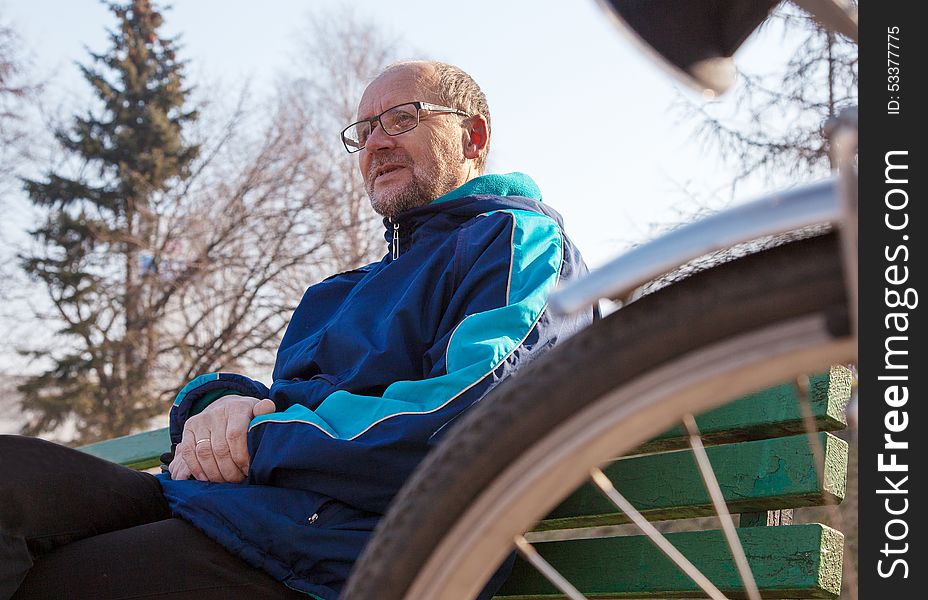 Eldely Man Sitting On A Bench Near His Bicycle In A City Park