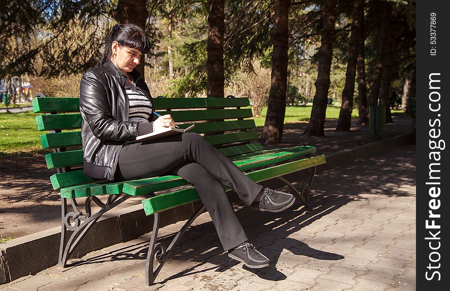 Young beautiful woman in a black leather jacket and black pants sitting on a park bench writing a pen in notebook on sunny spring day. Young beautiful woman in a black leather jacket and black pants sitting on a park bench writing a pen in notebook on sunny spring day