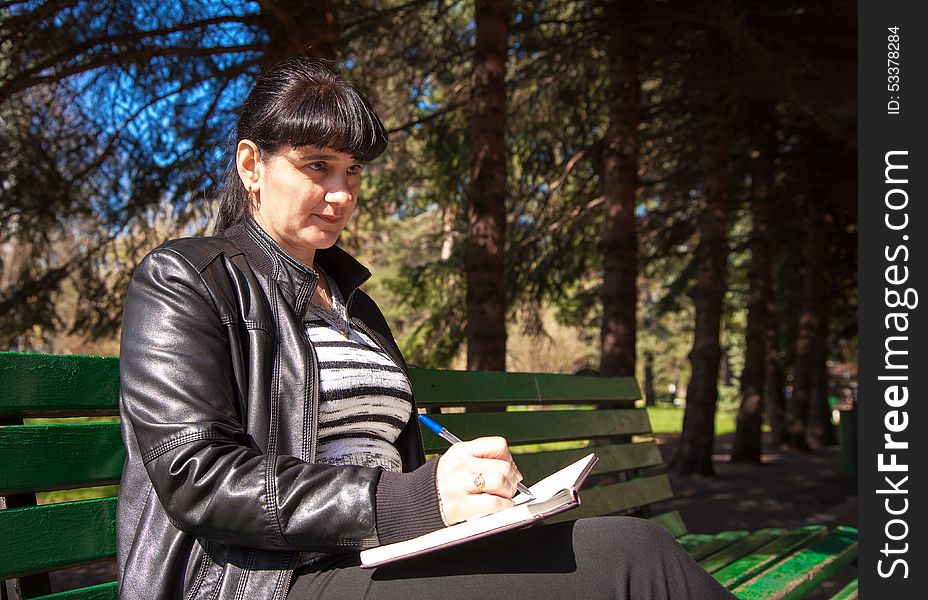 Young beautiful woman in a black leather jacket and black pants sitting on a park bench writing a pen in notebook. Young beautiful woman in a black leather jacket and black pants sitting on a park bench writing a pen in notebook