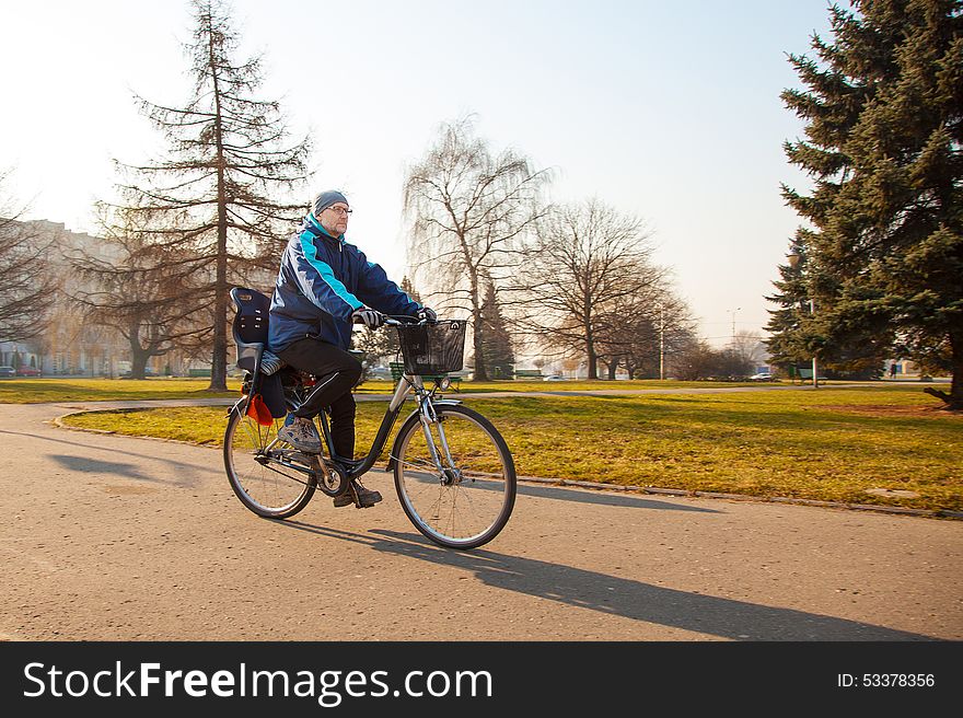 Elderly man wearing glasses, a blue jacket, black trousers and a gray hat, riding a bike on the sidewalk. Elderly man wearing glasses, a blue jacket, black trousers and a gray hat, riding a bike on the sidewalk