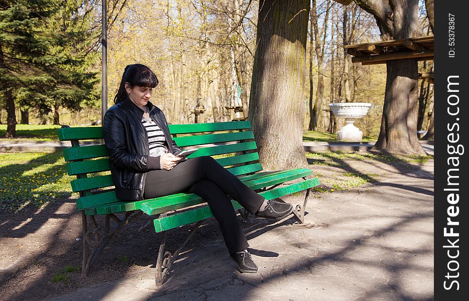 Young beautiful woman in a black leather jacket and black pants sitting on a park bench holding a tablet. Young beautiful woman in a black leather jacket and black pants sitting on a park bench holding a tablet