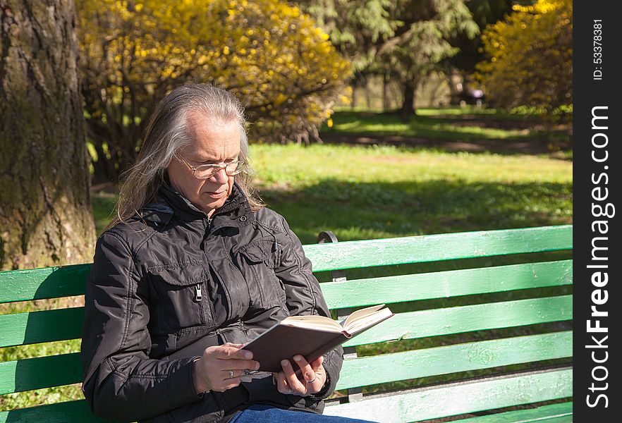 Haired Elderly Man Reading A Book Sitting On A Bench In City Par