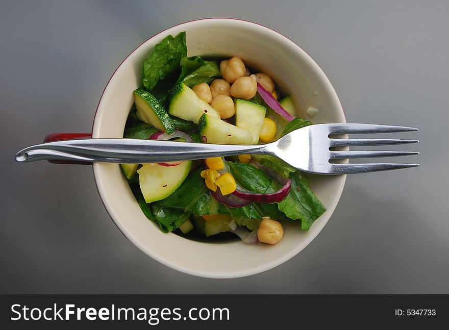 A summer salad in a ceramic bowl that includes, lettuce, garbanzo beans (chick peas), corn, onions, and zucchini. Shown from above with a fork.