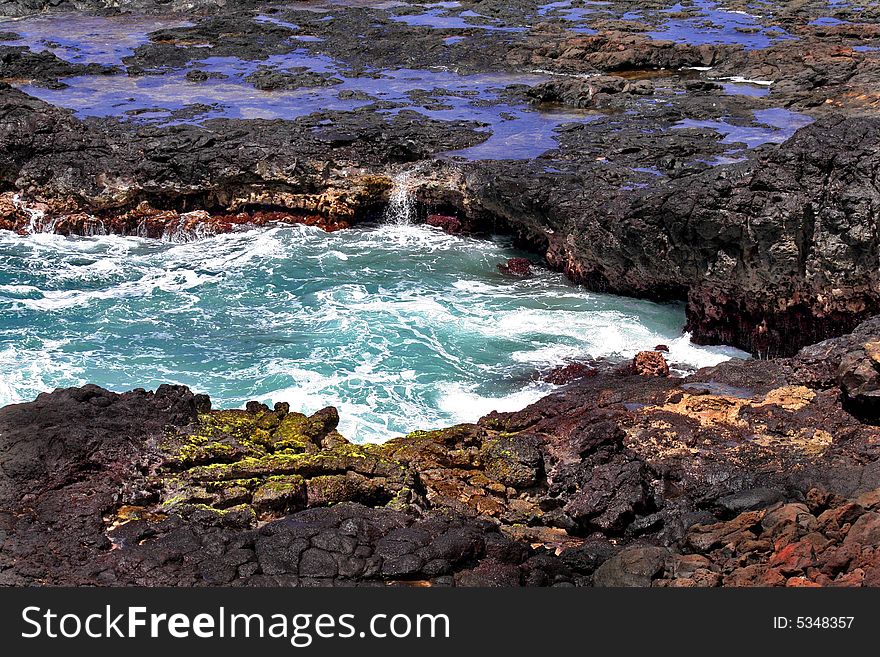 Ocean wave splashing in black lava rock on beach, blue pools of water in background, in foreground, green algae on black rock and some reddish rocks. Ocean wave splashing in black lava rock on beach, blue pools of water in background, in foreground, green algae on black rock and some reddish rocks