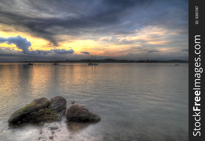 A cluster of rocks on a beach opposite a beacon and a moored yacht as seen at an overcast sunset. A cluster of rocks on a beach opposite a beacon and a moored yacht as seen at an overcast sunset