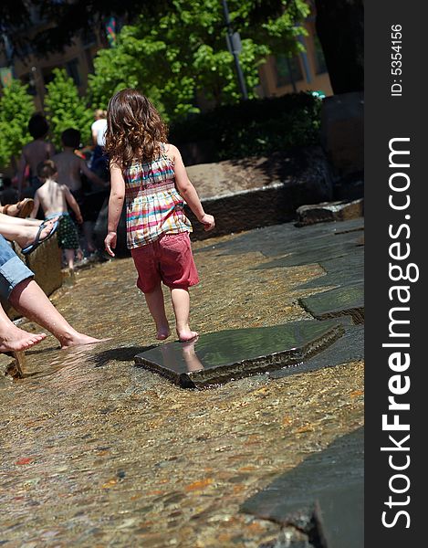 Cute young girl playing in a public fountain on a summer day. Vertically framed shot with the little girl facing away from the camera.