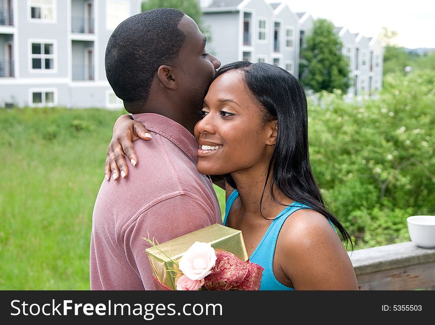 Attractive young woman hugging a man with one arm and holding a present in the other arm. Horizontally framed, outdoor shot on a deck, with the woman looking towards the camera at on off angle. Attractive young woman hugging a man with one arm and holding a present in the other arm. Horizontally framed, outdoor shot on a deck, with the woman looking towards the camera at on off angle.