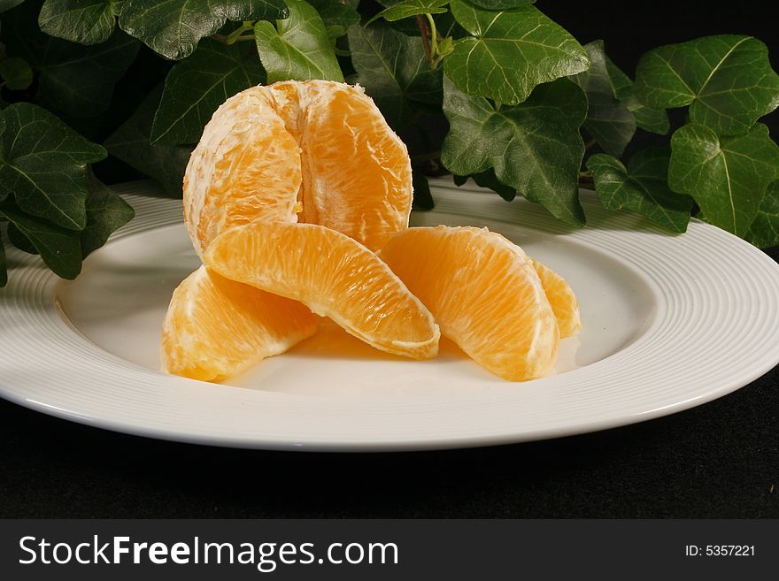 A display of a peeled orange with orange wedges on a white plate which is sitting on a black cloth.  There is ivy in the background.