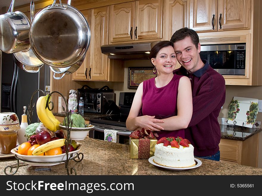Attractive couple hugging in the kitchen by a counter covered with a cake and a present. The pair are laughing heartily while looking at the camera. Horizontally framed shot. Attractive couple hugging in the kitchen by a counter covered with a cake and a present. The pair are laughing heartily while looking at the camera. Horizontally framed shot.