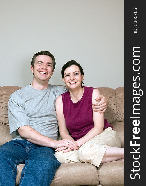 Attractive couple sitting on their living room couch with their arms around each other. Both are smiling at the camera. Vertically framed shot. Attractive couple sitting on their living room couch with their arms around each other. Both are smiling at the camera. Vertically framed shot.