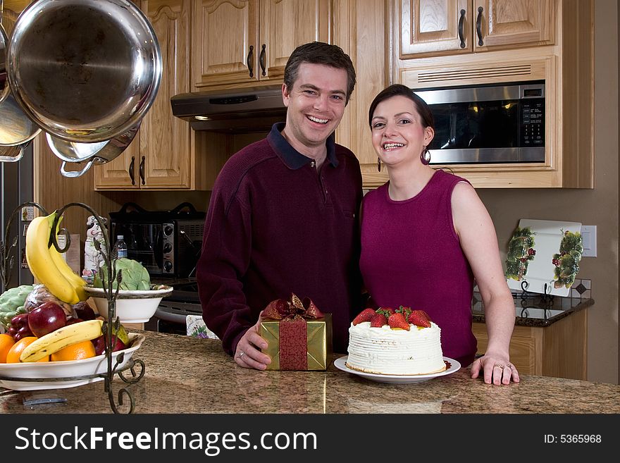 Attractive couple standing by a gift and a cake in their kitchen. Both are grinning widely and looking at the camera. Horizontally framed shot. Attractive couple standing by a gift and a cake in their kitchen. Both are grinning widely and looking at the camera. Horizontally framed shot.