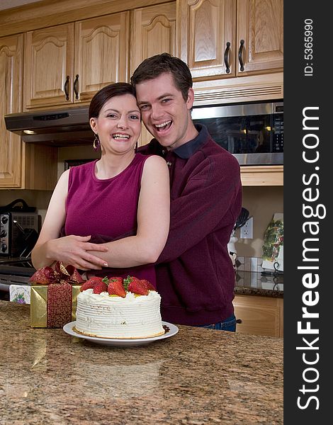 Attractive couple hugging in the kitchen by a counter covered with a cake and a present. Pair are laughing heartily and looking at the camera. Vertically framed shot. Attractive couple hugging in the kitchen by a counter covered with a cake and a present. Pair are laughing heartily and looking at the camera. Vertically framed shot.