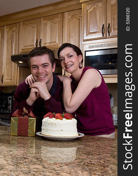 Attractive couple leaning on their kitchen counter which is covered with a cake and a present. They have a huge smile on their faces and are looking at the camera. Vertically framed shot. Attractive couple leaning on their kitchen counter which is covered with a cake and a present. They have a huge smile on their faces and are looking at the camera. Vertically framed shot.