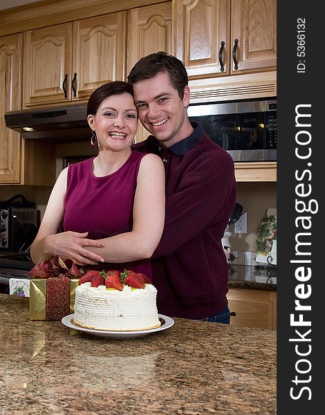 Attractive couple hugging in the kitchen by a counter covered with a cake and a present. Vertically framed shot. Pair are smiling at the camera. Attractive couple hugging in the kitchen by a counter covered with a cake and a present. Vertically framed shot. Pair are smiling at the camera.