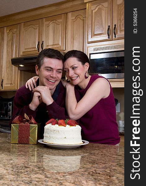 Attractive couple leaning on their kitchen counter which is covered with a cake and a present. They are laughing heartily and looking directly at each other. Vertically framed shot. Attractive couple leaning on their kitchen counter which is covered with a cake and a present. They are laughing heartily and looking directly at each other. Vertically framed shot.