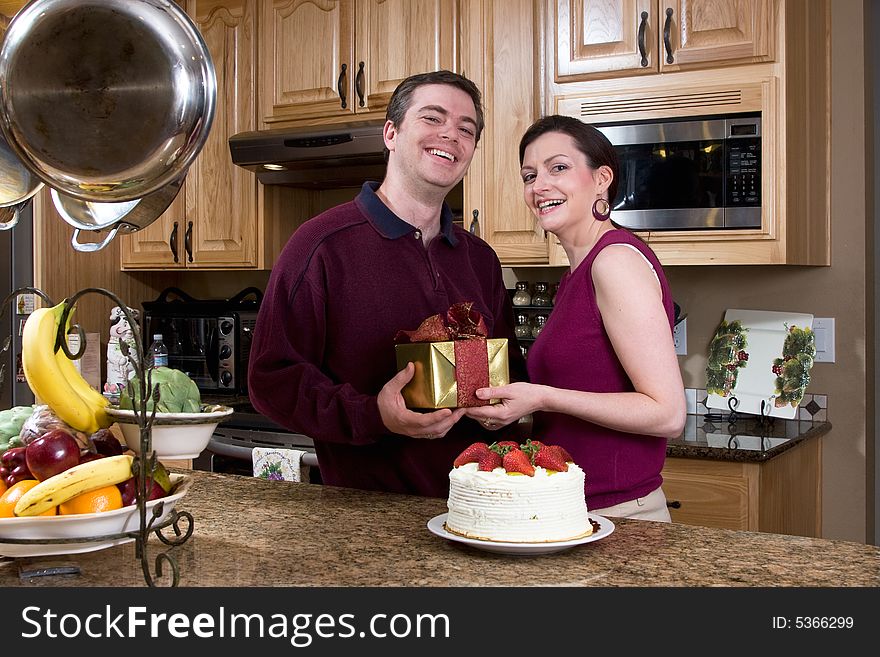 Attractive couple laughing at the camera while standing in the kitchen and exchanging gifts and cake. Horizontally framed shot. Attractive couple laughing at the camera while standing in the kitchen and exchanging gifts and cake. Horizontally framed shot.