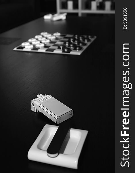 White ashtray, pack of cigarettes, box of matches and chessboard on a black table in an home interior. White ashtray, pack of cigarettes, box of matches and chessboard on a black table in an home interior.