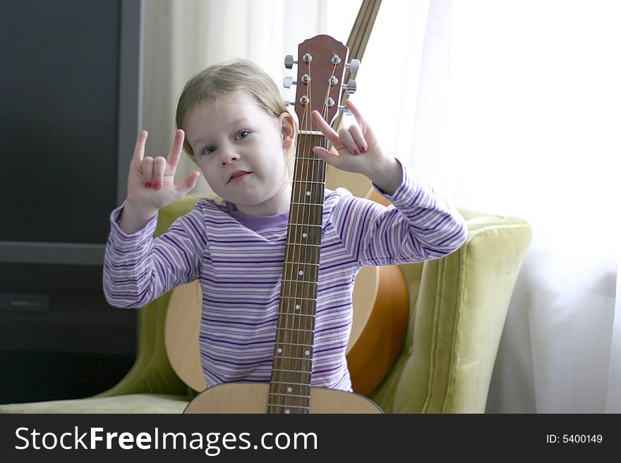 Female child age six holding an acoustic guitar and gesturing the rock on sign with her hands. The child is wearing a purple striped shirt and sitting on a green chair. Female child age six holding an acoustic guitar and gesturing the rock on sign with her hands. The child is wearing a purple striped shirt and sitting on a green chair.