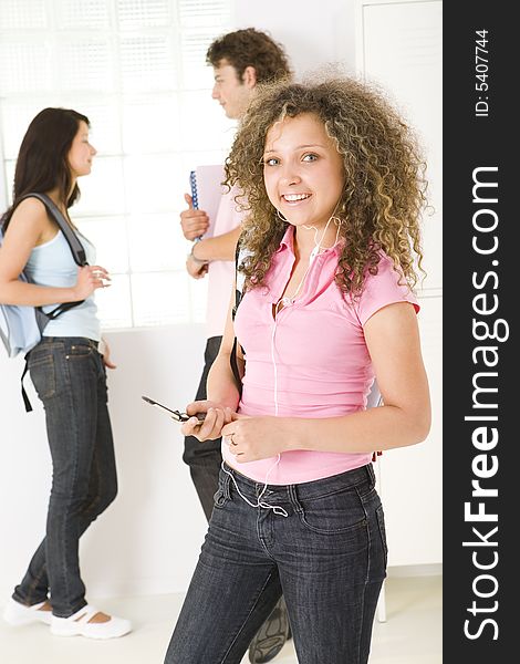 Three schoolmate standing near window. A boy holding notebooks and talking with girl in blue shirt. A girl in ping shirt smiling and listening to music by headset. Focused on girl in pink shirt. Three schoolmate standing near window. A boy holding notebooks and talking with girl in blue shirt. A girl in ping shirt smiling and listening to music by headset. Focused on girl in pink shirt.