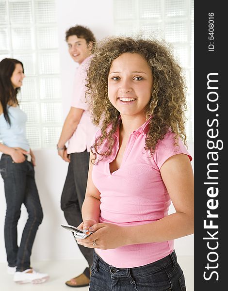 Three happy friends standing near window. A boy talking with girl in blue shirt. A girl in pink shirt holding mobile phone and looking at camera. Focused on girl in pink shirt. Three happy friends standing near window. A boy talking with girl in blue shirt. A girl in pink shirt holding mobile phone and looking at camera. Focused on girl in pink shirt.