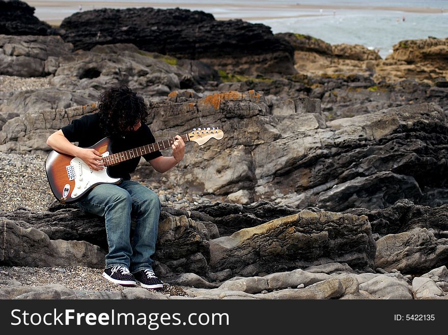 A teenage boy playing the guitar on the rocks near the beach. A teenage boy playing the guitar on the rocks near the beach.