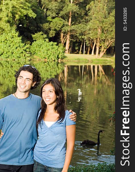 Happy, smiling couple embrace as they stand in front of a wooded pond with swans in it. Vertically framed photograph. Happy, smiling couple embrace as they stand in front of a wooded pond with swans in it. Vertically framed photograph.
