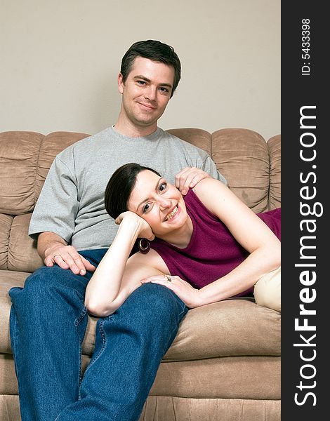 Smiling, happy couple sitting on a couch. They are smiling and he has his arm around her as she lays across his lap. Vertically framed photograph. Smiling, happy couple sitting on a couch. They are smiling and he has his arm around her as she lays across his lap. Vertically framed photograph